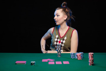 Emotional young lady in a white blouse drinking wine from a glass and playing cards on a table on green cloth in a casino