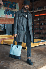 Wall Mural - Fashionable young man in black waxed trench coat, bucket hat and with blue bag, walking in loft interior room with painting and chain link.