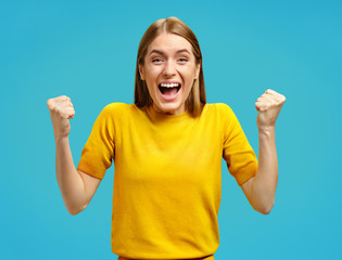 Wall Mural - Excited girl with widely open mouth holds hands clenched in fists, exclaimed with positiveness. Photo of attractive girl in yellow sweater on blue background. Emotions and pleasant feelings concept.