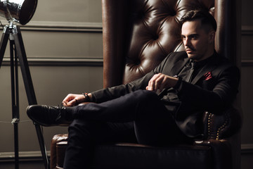 Wall Mural - Young handsome man posing for a fashion shoot in a studio. Fashion as a lifestyle. Man wearing a beard. Model sitting on a sofa in tuxedo. Successful fashionable businessman. Business look advertising