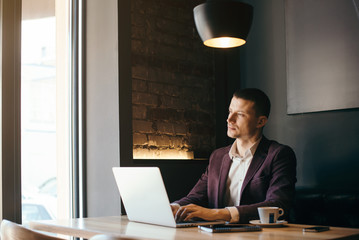 Young handsome man sitting in office with cup of coffee and working on project connected with modern cyber technologies. Businessman with notebook trying to keep deadline in digital marketing sphere.