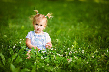 Happy funny blonde baby girl with two little braids in white t-shirt and jeans bodykit sitting on green grass background