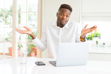 Poster - African american business man working using laptop clueless and confused expression with arms and hands raised. Doubt concept.