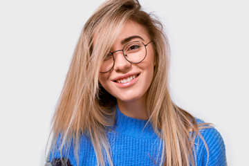 Pretty positive European young woman in stylish loose sweater and round transparent eyewear, smiling broadly and looking to the camera, isolated over white background. People emotion