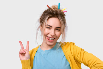 Joyful young woman showing tongue and peace gesture while looking to the camera and standing against white background. Happy female wearing casual outfit and colorful markers on the hair having fun