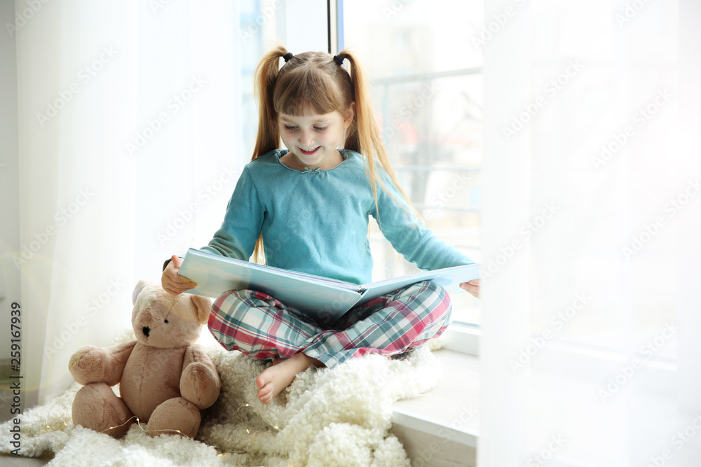 Cute little girl with teddy bear sitting near window and reading book ...