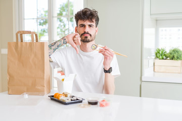 Sticker - Young man eating asian sushi from home delivery with angry face, negative sign showing dislike with thumbs down, rejection concept