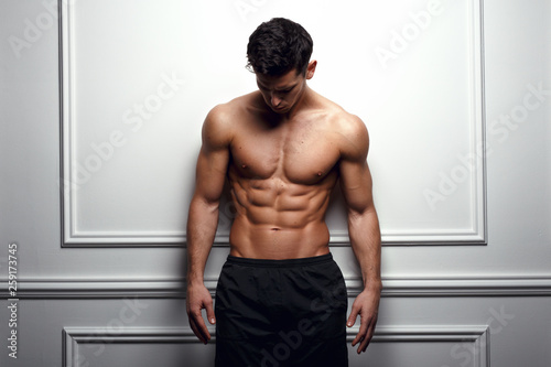 Cuadros en Lienzo Athlete, muscular man at the white wall poses shirtless, showing six pack abs, white background