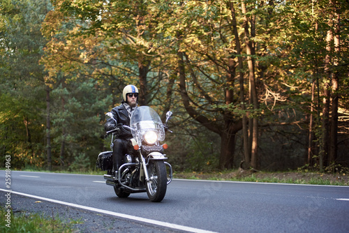 Cuadros en Lienzo Handsome bearded biker in sunglasses, helmet and black leather clothing riding cruiser motorcycle along asphalt road winding among tall green trees on sunny summer evening