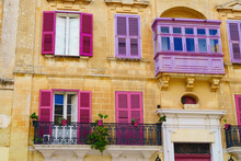 Facade Of The House With Purple And Pink Maltese Balconies, Plants In Pots , Windows With Shutters On Yellow Limestone Townhouse. Typical Street Scene In Ancient City Of Mdina, Malta.