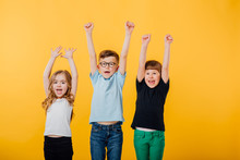 Three Beautiful Children, A Little Boy And Two Little Girls Jumping Up, Their Hands Raised, Happy, Glad, Isolated Yellow Background, Copy Space