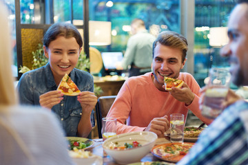 Cheerful young people enjoying eating delicious pizza and talking during dinner with friends in restaurant