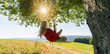Unrecognizable girl in white dress swaying on a tree swing on peaceful evening. Lady sitting on a wooden swing and looking at golden sunset. Young woman swinging at sunrise