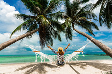 Happy Beautiful Girl In A Straw Hat And Shorts, Sitting Back On A Beach Hammock, Arms Raised To The Top, Between Two Palm Trees, On The Seashore Of A Tropical Island, Traveling Summer Vacation