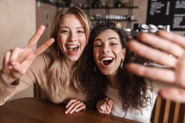 Canvas Print - Excited happy pretty girls friends sitting in cafe take a selfie by camera.