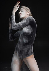 Beautiful slim girl wearing a black gymnastic bodysuit covered with white powder talcum dust jumps dances on a dark. Artistic conceptual and advertising photo.