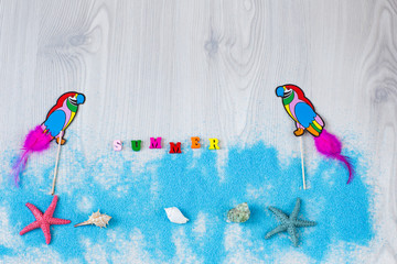 on a light background  blue decorative sand, two parrots, starfish, shells and the word summer written in wooden letters