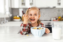 Beautiful Little Girl Having Breakfast With Cereal, Milk And Blueberry In Kitchen
