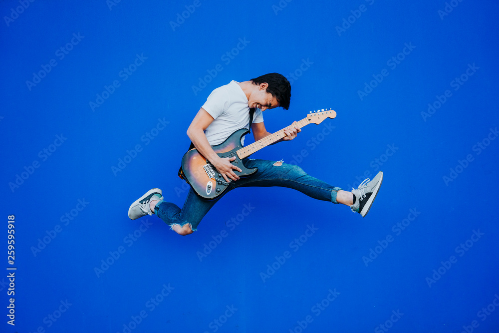 young man jumping with electric guitar on blue background - obrazy, fototapety, plakaty 