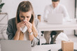 blonde businesswoman sneezing in tissue near coworker in office