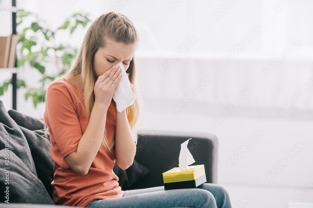 attractive blonde woman sneezing while sitting on sofa with tissue box - obrazy, fototapety, plakaty 