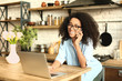 Female African-American freelancer talking by phone while working on laptop in kitchen