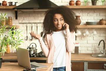 Wall Mural - Female African-American freelancer talking by phone while working on laptop in kitchen