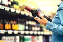 Alcohol Shelf In Liquor Store Or Supermarket. Woman Buying A Bottle Of Red Wine And Looking At Alcoholic Drinks In Shop. Happy Female Customer Choosing Merlot Or Sangiovese. Shopping Spirits Concept.
