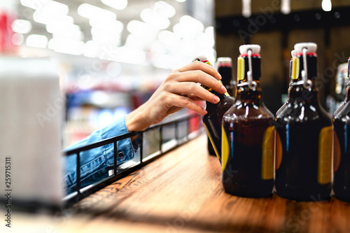 Customer taking bottle of beer from shelf in liquor store. Woman shopping alcohol or supermarket staff filling and stocking shelves in drink aisle. Cider, lager or dark. Retail worker doing inventory.