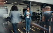 © JonoErasmus - Beautiful black african business woman standing in a busy lobby of an office looking down at a tablet with people walking past in a blur