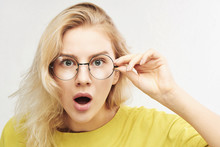 Surprised Woman In Round Glasses With Open Mouth And Bulging Eyes Looks Into Camera And Sees Something Incredible And Amazing Isolated On White Background