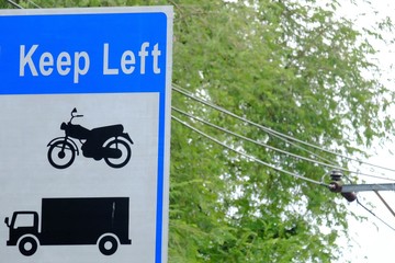 The pictures of motorcycle and a truck on a white blue traffic board with a word  Keep Left in the high view,green trees and electricity lines background 