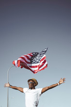 Excited Black Man In Sunglasses And Hat Holding Hands Apart With American Flag Flying Above Head On Blue Sky