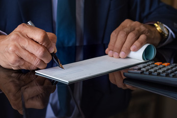 Wall Mural - Businessman  in black suit with necktie, hand writing and signing checkbook on the wooden table at office. Paycheck or payment by cheque concept.