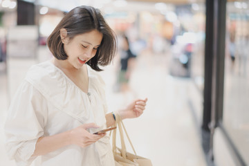 Young Asian beautiful woman shot hair shopping in a shopping mall store. Woman standing and holding a shopping bag and looking to her mobile phone in the store near big glass window.