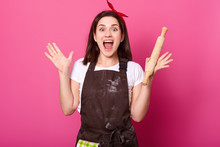 Baker Young Woman Puts Her Hands Up Holding Rolling Pin, Wears Brown Apron, White Tshirt, Opens Her Mouth Widely. Adorable Cute Female Is In High Spirits While Cooking New Dishes. Cook Concept.