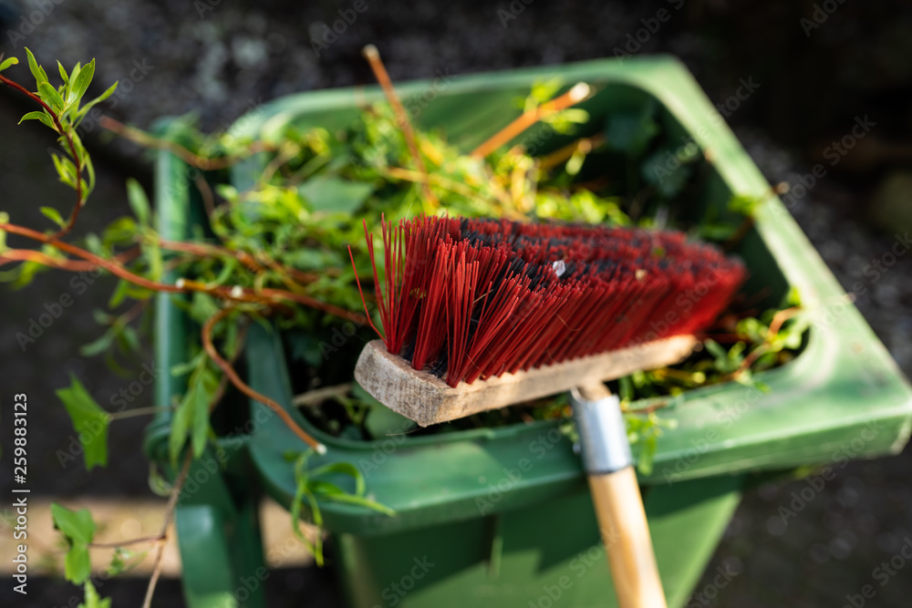 Green wheelie bin / garden waste container and broom filled with fruit and vegetable waste, garden waste, organic waste for composting and fermentation. Recycling garbage for a better environment.