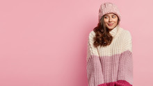 Beautiful Cheerful Woman With Charming Smile, Wears Knitted Sweater And Hat, Looks With Dreamy Satisfied Expression, Stands Against Pink Background, Blank Space For Your Advertisement Or Promotion