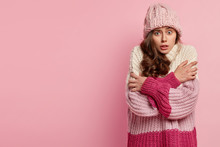 Worried European Woman Trembles With Fright, Crosses Hands Over Chest, Doesnt Likes Freezing Chilling Weather, Wears Warm Hat And Knitted Oversized Jumper, Stands Over Pink Wall With Empty Space