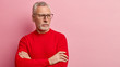 Retired bearded old man keeps arms folded, focused away with pensive look, wears spectacles and red jumper, stands over pink background, free space for your advertisement. People, age, thoughts