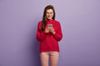 © wayhome.studio  - Photo of smiling Caucasian teenage girl focused in modern cell phone, enjoys online communication, wears long knitted red sweater, sends text messages to friend, stands over purple background