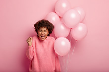 Half Length Shot Of Optimistic Overjoyed Female With Afro Haircut Clenches Fist, Celebrates Triumph, Graduation From University, Invites Friend On Party, Holds Bunch Of Pink Balloons, Exclaims Happily