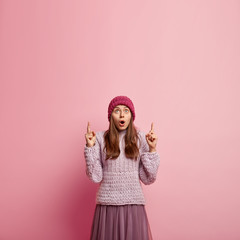 Wall Mural - Shocked beautiful woman with widely opened mouth, dressed in hat, knitted jumper and long skirt, points above at blank space for your advertising content. People, astonishment, advertisement