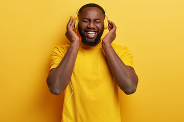 Wall Mural - Monochrome shot of overjoyed pleased Afro American man enjoys perfect loud sound in new headphones, dressed in yellow t shirt, has free time, entertains himeslf with music. Happy expression.