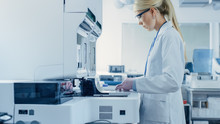 Female Research Scientist Putting Test Tubes With Blood Samples Into Analyzer Medical Machine. Scientist Works With Modern Medical Equipment In Pharmaceutical Laboratory.