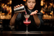 Professional female bartender pouring a smooth crimson cocktail through the sieve to the glass