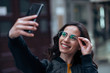 © bnenin - Headshot of a smiling young woman with eyeglasses taking selfie in the city.