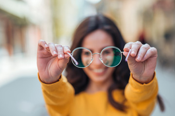 Young woman holding eyeglasses toward camera outdoors.