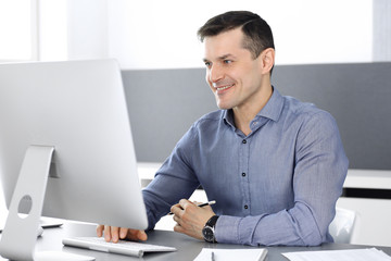 Cheerful smiling businessman working with computer in modern office. Headshot of male entrepreneur or director of a company at the workplace. Business concept 