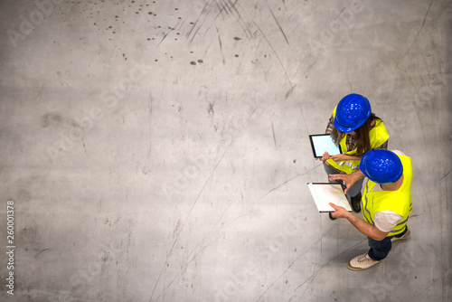 Fotomural Top view of two industrial workers wearing hardhats and reflective jackets holding tablet and checklist on gray concrete background